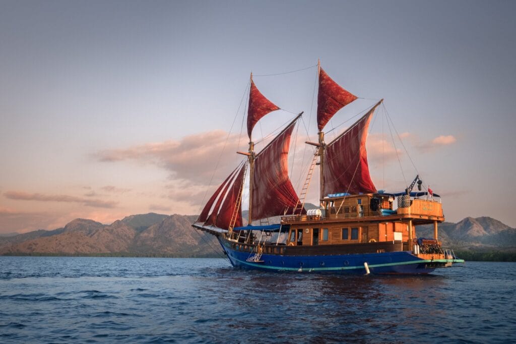 Tiger Blue 1 broadside under red sails at dusk, varnished two-deck wooden cabin, Komodo mountains behind