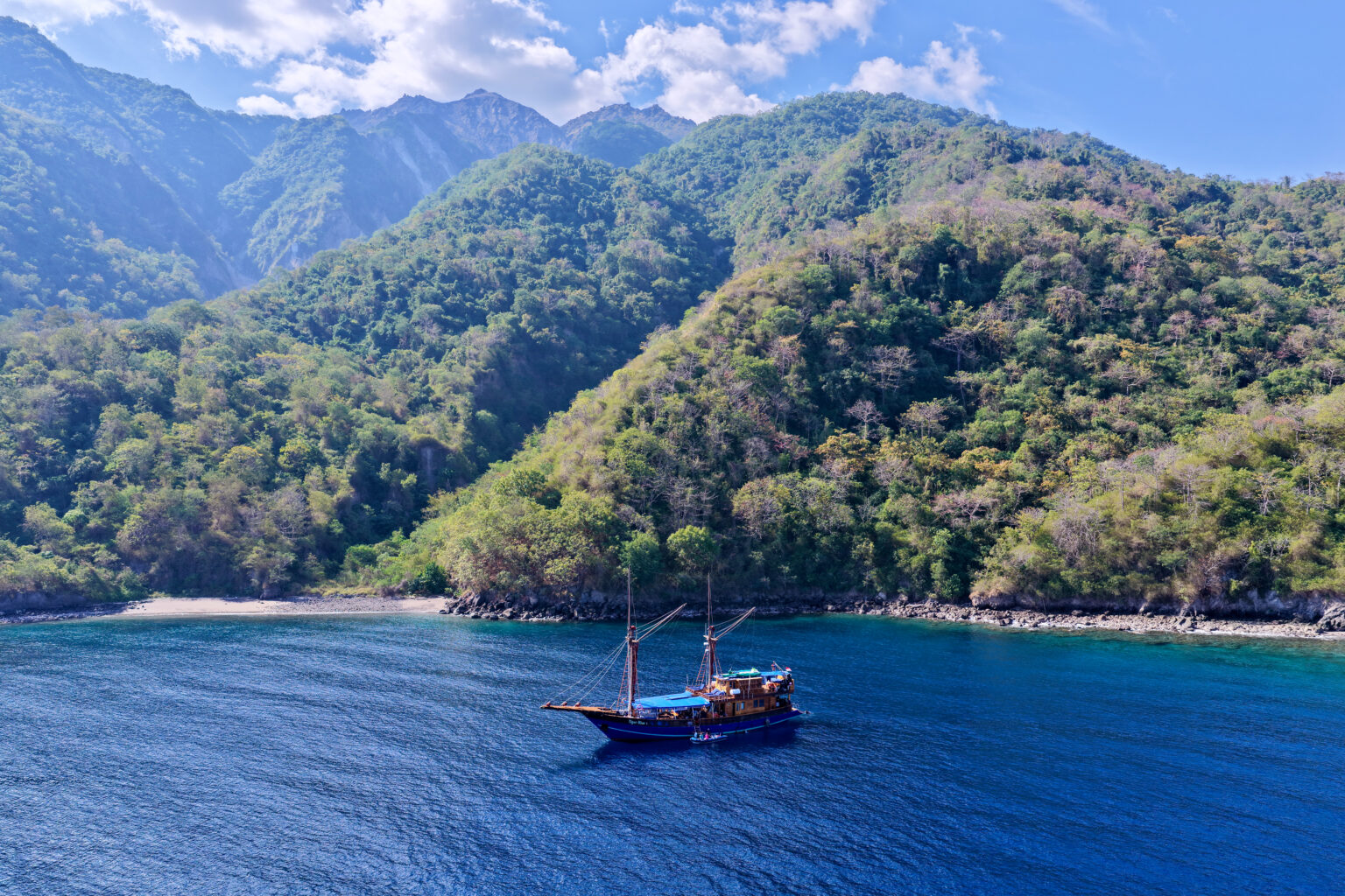 Tiger Blue at anchor in a sheltered bay below forested mountains, seen from a drone