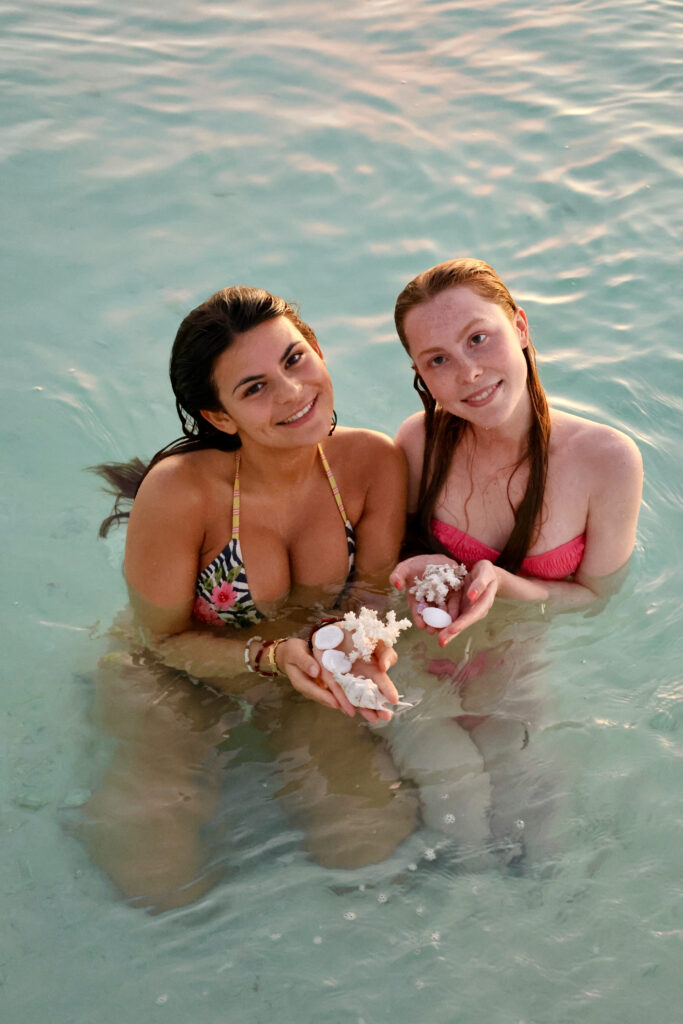 Two guests smiling in clear shallow water, holding shells they have found