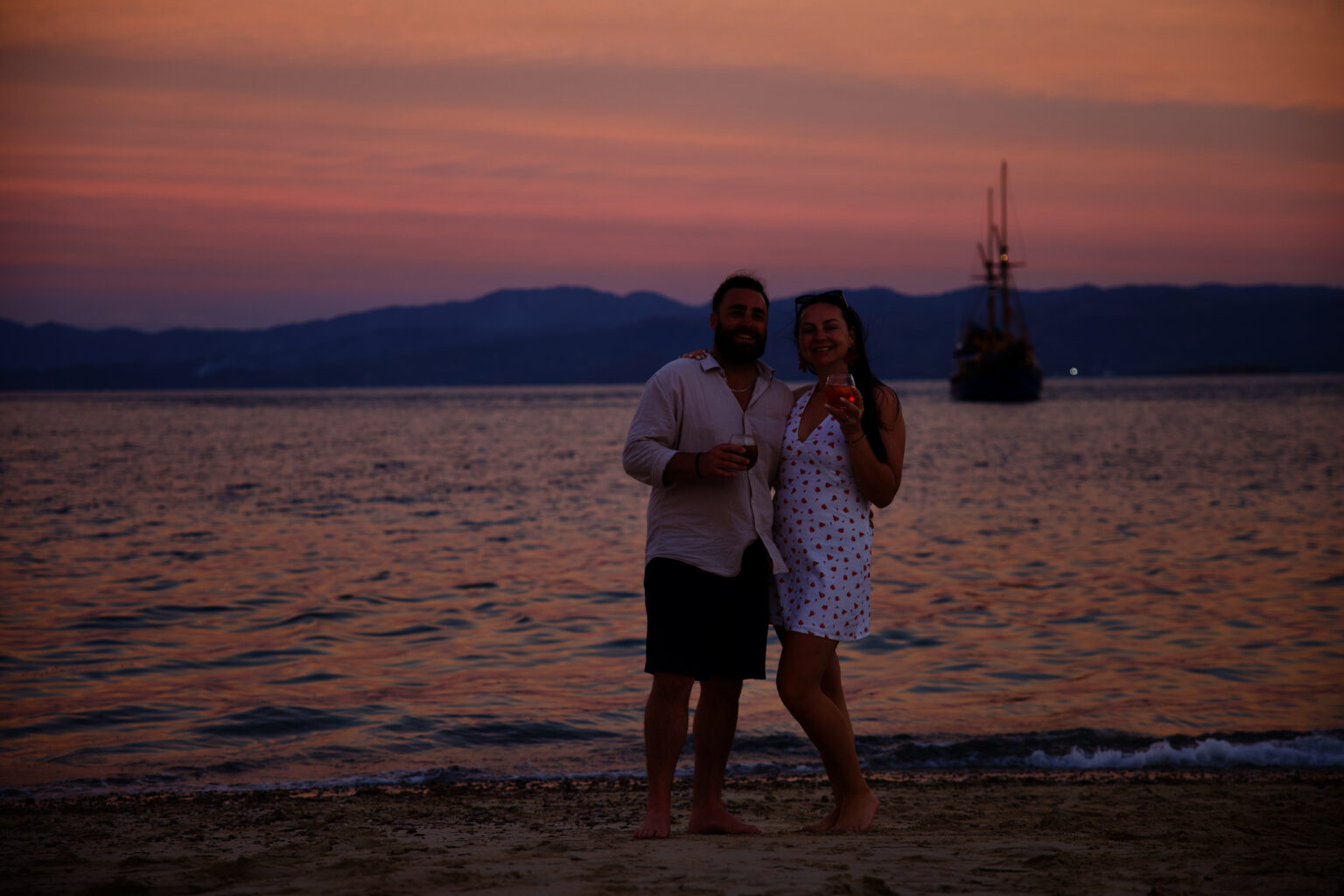Two guests with drinks on the beach at sunset, with Tiger Blue at anchor behind them