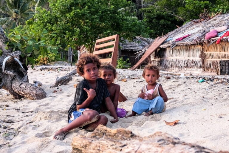 Village children sitting on a sandy beach beside thatched huts